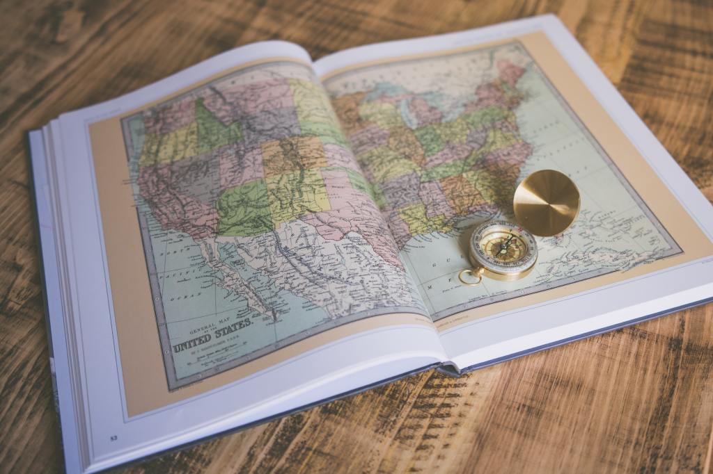 A book lies open on a wooden table, turned to map of the United States of America that spans two pages. A golden compass sits open on the right-hand page.