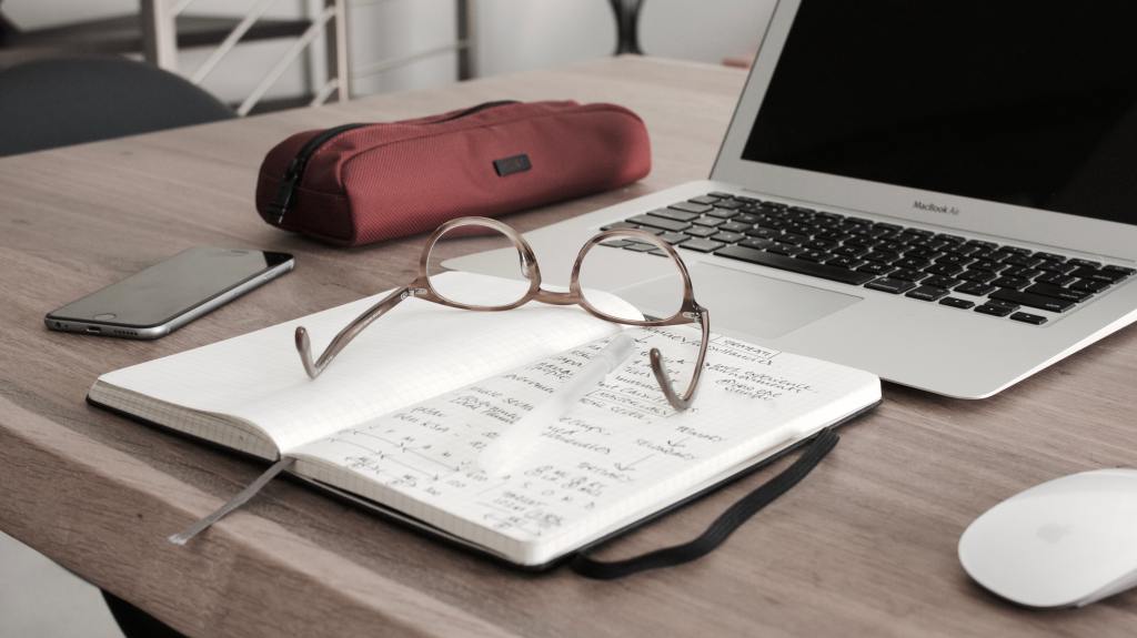 A laptop on a table next to a red pencil bag, a smart phone and a journal that is lying open with a pair of glasses sitting on top. The laptop screen is blank.
