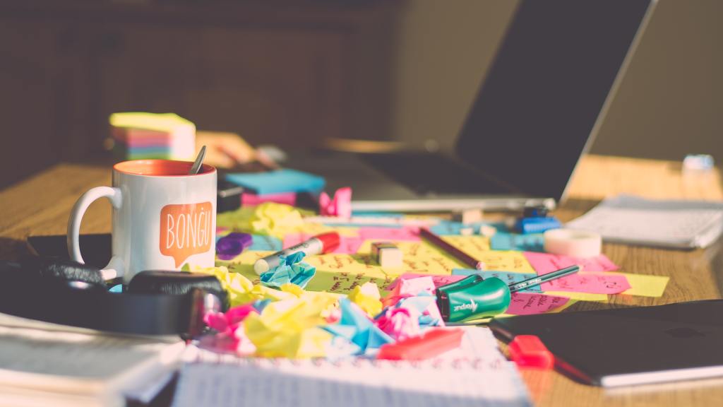 Picture of a messy desk. Laptop is open in the back, crumpled sticky notes are scattered across the desk, a half-empty coffee mug and various other items.