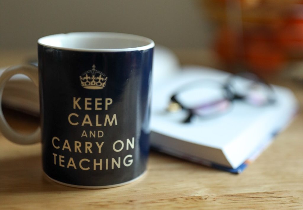 A blue mug that has an icon of a crown and says 'Keep Calm and Carry On Teaching' sits in front of an open book. A pair of glass sits on top of the book.
