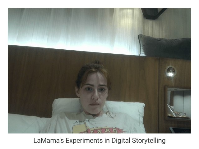 A webcam capture of a woman with red hair in a bun. She looks ready to go to bed and is sitting in what appears to be a hotel bed. You can see just the headboard behind her and a few pillows. The caption of the capture is 'LaMama's Experiments in Digital Storytelling'.