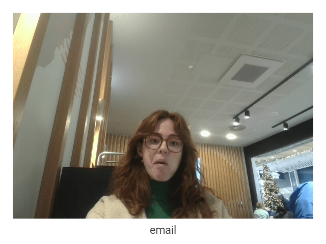 A webcam capture of a woman with curly short red hair wearing glasses, a beige cardigan, and green turtleneck. In the background is a seating area in a cafe, and you can make out a Christmas tree in the distance to the right of the seating area outside the cafe. The caption of the capture is 'email'.