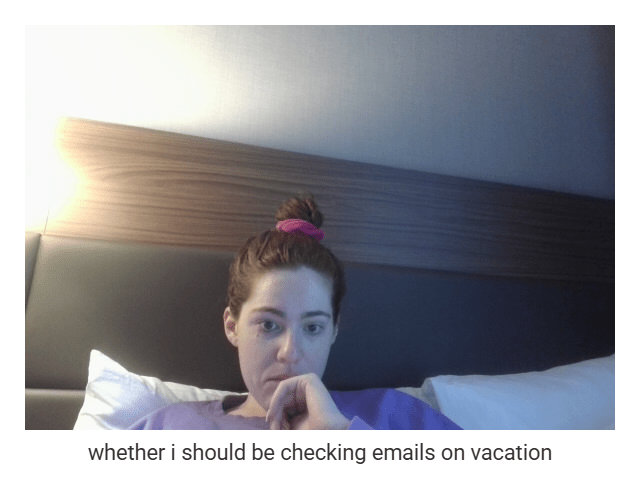 A webcam capture of a woman with red hair up in a high bun with a hot pink scrunchy. She is wearing a big purple sweatshirt. Behind her is the headboard of a hotel bed. The caption of the capture is 'whether i should be checking my emails on vacation'.