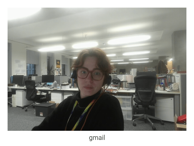 A webcam capture of a woman in an empty office. She has curly red hair, in a bun, with glasses. She has headphones over her ear and a rainbow University of Edinburgh lanyard around her neck. The caption of the capture is 'gmail'.