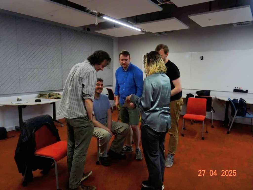 An enclosed rehearsal space. A woman speaks to 4 other male actors, either sitting or standing in a semi-circle towards her.