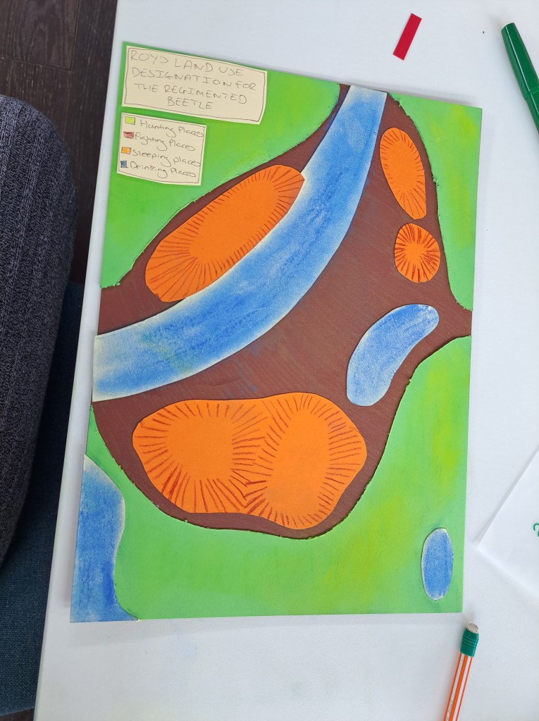 An artwork with colourful paper cut outs depicting a map of 'Roy's Land Use Designation for the Regimented Beetle'. The map has a colour-coded key with categories such as 'hunting places', 'fighting places', 'sleeping places', and 'drinking places'. The map appears to be an organic collection of various shapes and colours.