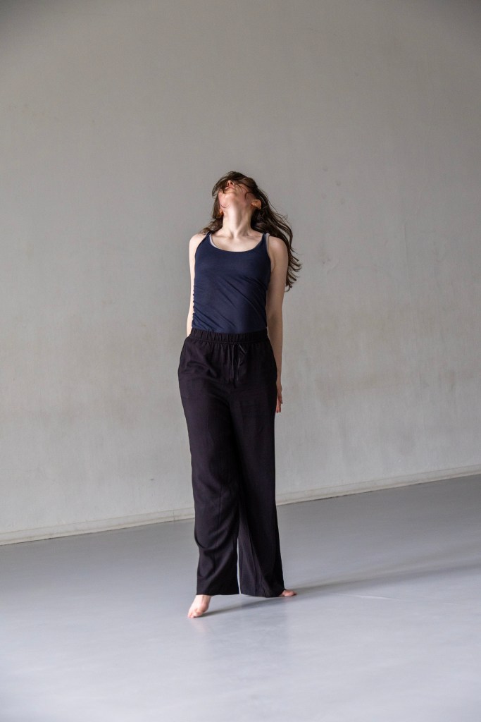 A dancer against a light grey concrete wall. She has long dark hair, a navy tank top, black joggers, and bare feet. She leans her head back, pushes her shoulders back, and hides her hands from view of the camera.