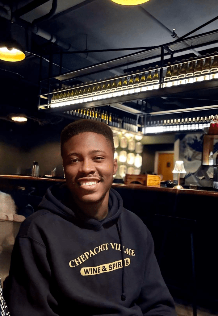 A picture of a black man with a shaved haircut, smiling in a black hoodie. There is a restaurant/bar in the background.