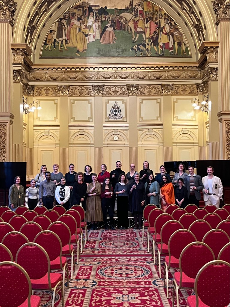 A group of people posing for a photo behind rows of empty red chairs placed in a regal, Regency Era hall.