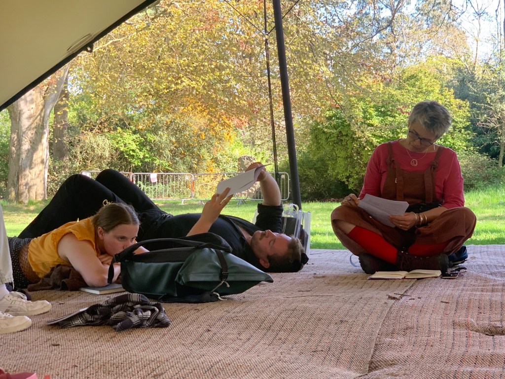 A man laying down reading outside, covered by a tent. Others are sitting and laying in close proximity and also look to be reading intently