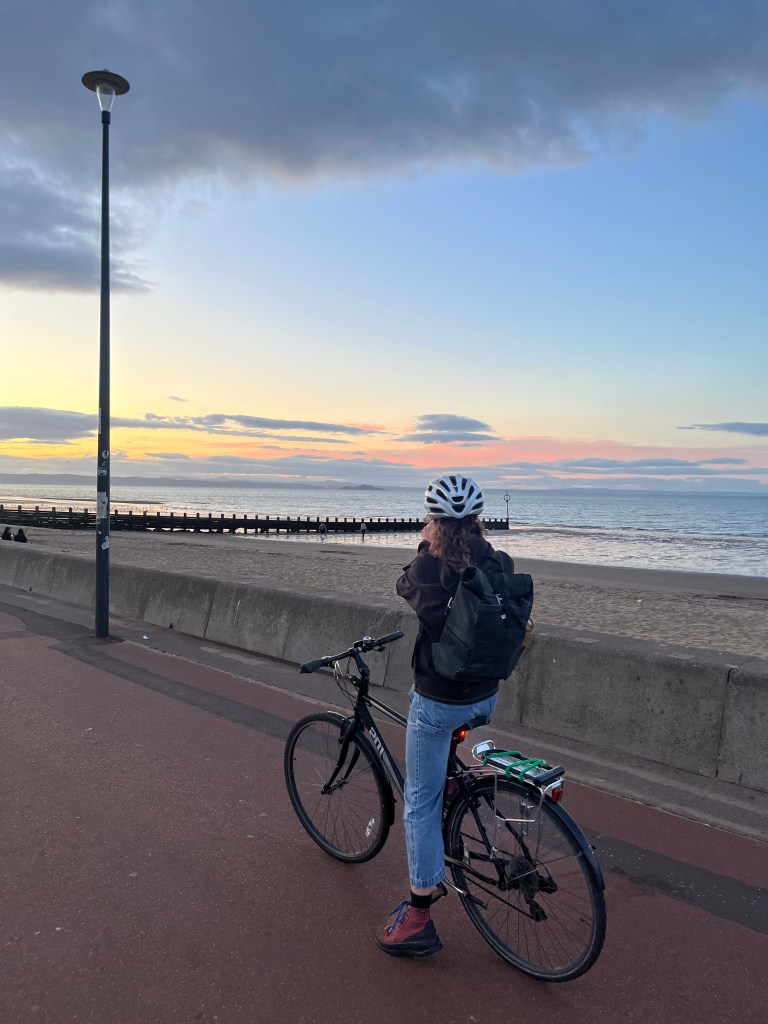 A cyclist on a beach promenade, stopping to take a photo of the sun setting over the shore.