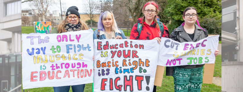 A group of four protesters hold up hand drawn signs voicing opposition to redundancies and salary cuts at their university