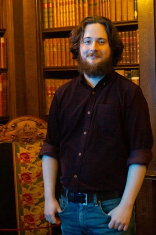 A man with longish auburn hair and a full beard in a button down, dark coloured, long sleeve shirt and blue jeans. He is standing in what appears to be an old library, with an old-looking bookcase behind him.