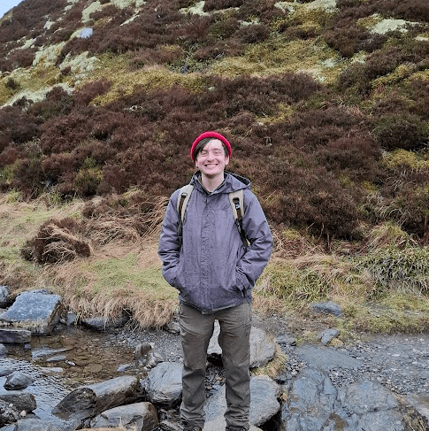 A person standing in a small rivulet dotted with rocks. A stunning, grassy and hilly landscape is behind them. They are wearing outdoor apparel, including a rain jacket, backpack, and red beanie.
