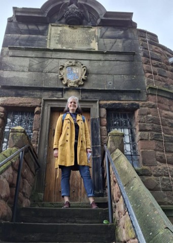 A woman with a greying bob wearing a yellow raincoat/trench coat, jeans, sensible walking shoes, and a backpack. She is standing in front of what appears to be a historical stone building with a crest just above the doorway and an inscription just above the crest.