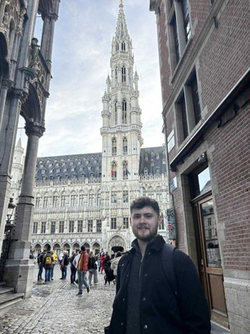 A man with short dark brown hair and a full beard, wearing a dark-coloured, light winter coat with a zipped up jumper on underneath. Behind him is what appears to be a European cathedral with a very tall, pointy tower. In the background, there are tourists milling about in what appears to be a European piazza/square.