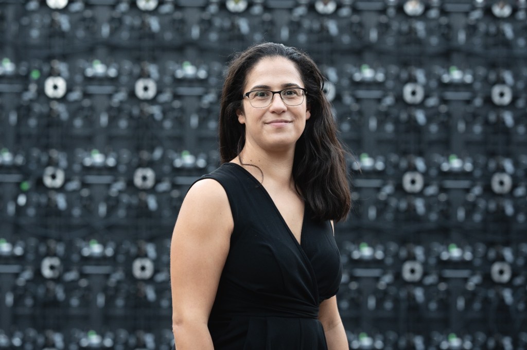 A headshot of a woman with long dark hair, glasses, and a Mediterrannean complexion. She is wearing a fitted black top and is against a blurred, patterned background.