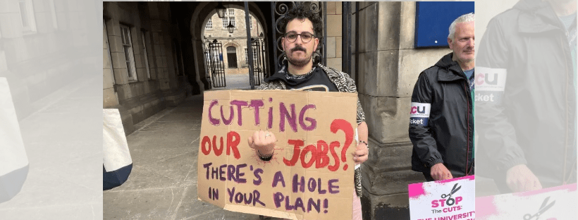 An image of a protester with a stone archway in the background. He is holding a sign that reads 'Cutting our jobs? There's a hole in your plan'! with a hole through the middle of the sign and his fist through the hole.