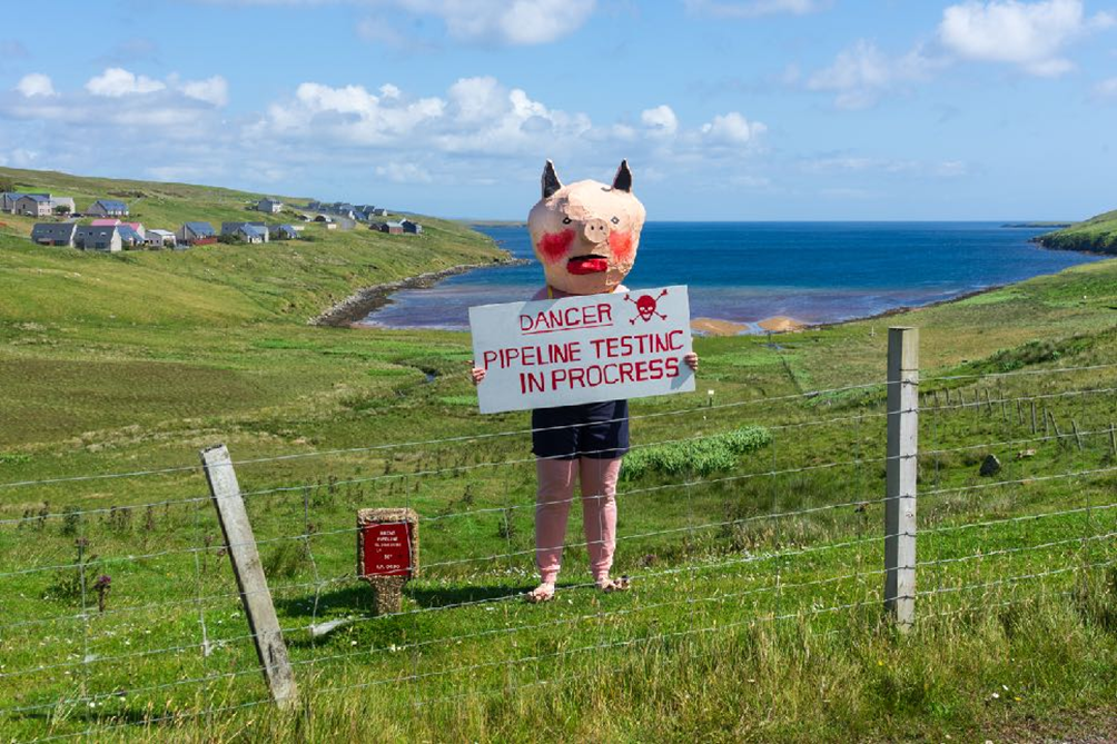 Re-enactment of Up Helly Aa skit ‘BP Pigs’ (1979) on top of the Brae Field oil pipeline. Following the pipeline up from the sea towards the Sullom Voe oil terminal leads Piggie to a little house from which oil and gas workers can access the Brae pipeline for maintenance and cleaning purposes. Part of The Moving Energy Theatre tour, 2025.
