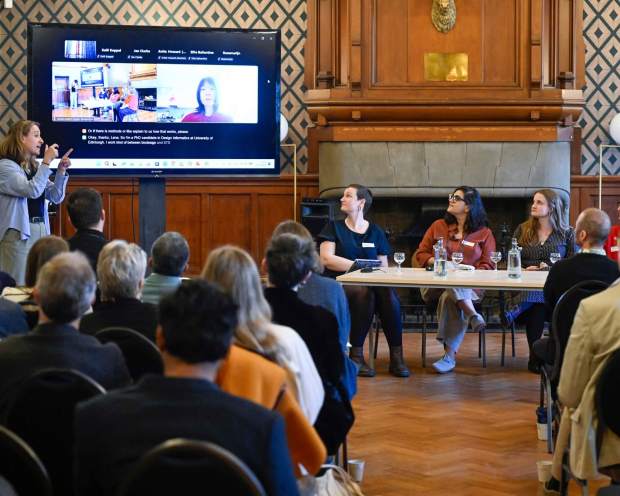 BSL interpreter presenting beside a screen showing a hybrid panel, with seated panellists listening and an audience in the foreground.