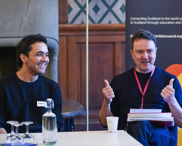 Two panel speakers seated at a table, one gesturing with both thumbs while speaking, with papers, water glasses, and a British Council banner behind them.