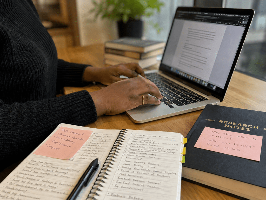 A doctoral researcher typing on a laptop beside handwritten notes and a PhD proposal notebook.