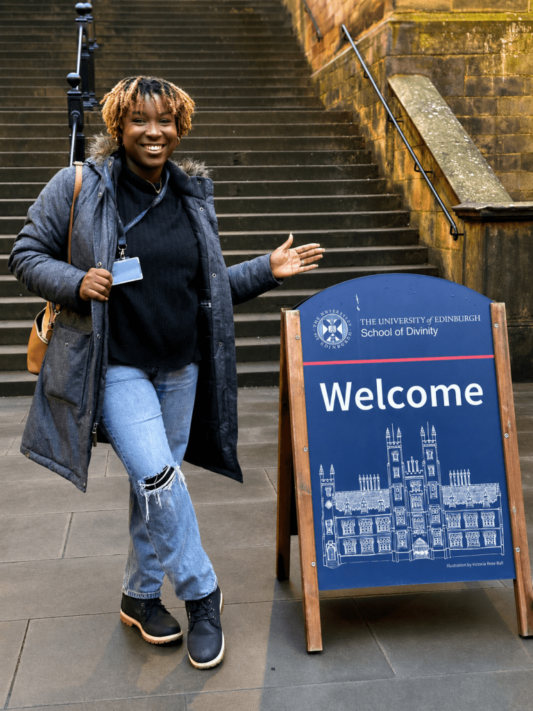 Olivia Shaw-Lovell standing beside a University of Edinburgh School of Divinity welcome sign.