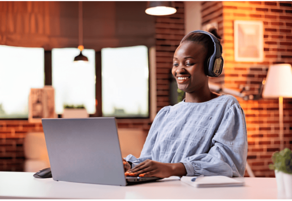 Smiling Black woman wearing headphones while working on a laptop at a desk in a warmly lit room.