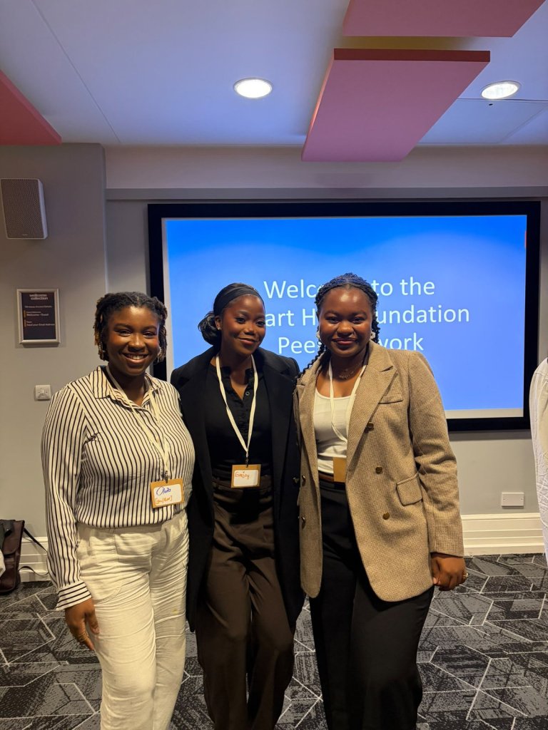 Three Black women smiling together at a Stuart Hall Foundation Peer Network gathering, standing in front of a blue welcome presentation screen at the Wellcome Collection in London.