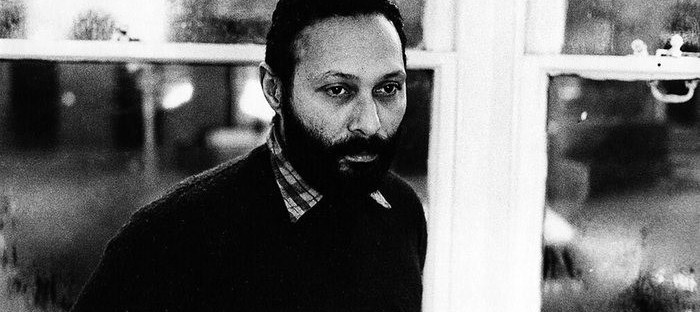 Black-and-white portrait of cultural theorist Stuart Hall, wearing a dark jacket over a checked shirt, seated in front of a window and looking thoughtfully to one side.