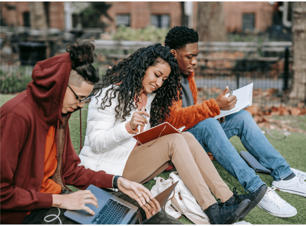 Three university students sitting together on grass outdoors, studying with a laptop and notebooks on a university campus.
