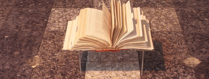 An open book with pages fanned upwards rests on a transparent display stand above a floor covered in patterned brown tiles.