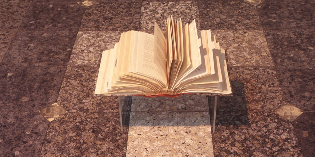 An open book with pages fanned upwards rests on a transparent display stand above a floor covered in patterned brown tiles.
