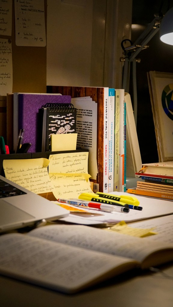 Late-night study desk with open books, handwritten notes, sticky notes, stationery and a laptop.